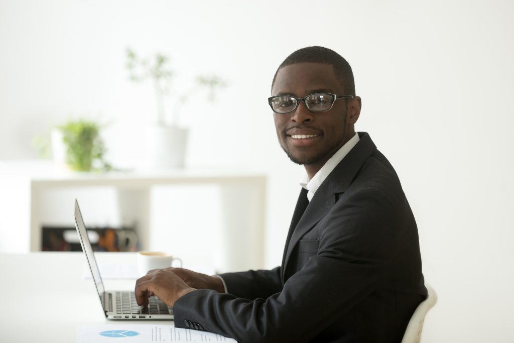 African-american smiling businessman in suit and glasses working on laptop, friendly black financial accountant, marketing manager, business analyst or ceo looking at camera at workplace, portrait